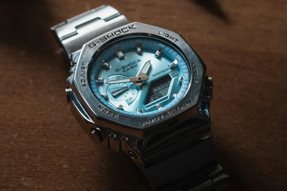Vollmetall-G-Shock liegt schräg auf einer Holzfläche, gebürstete Bandglieder und polierte Lünettenfacetten wechseln sich im warmen Licht ab
