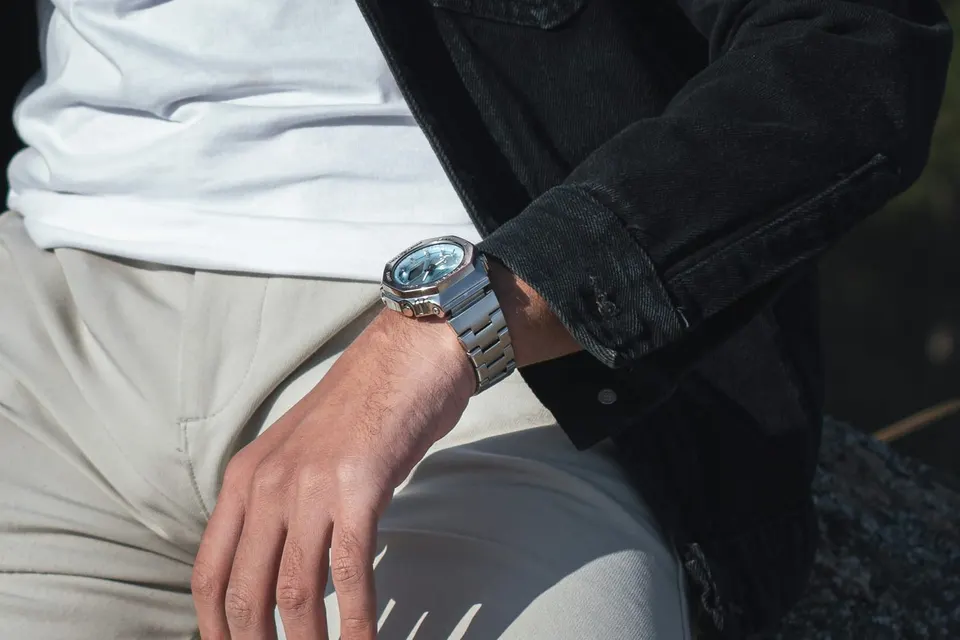 Mann in schwarzer Jeansjacke und heller Hose sitzt auf einem Felsen, am Handgelenk eine Metall-G-Shock mit hellblauem Zifferblatt