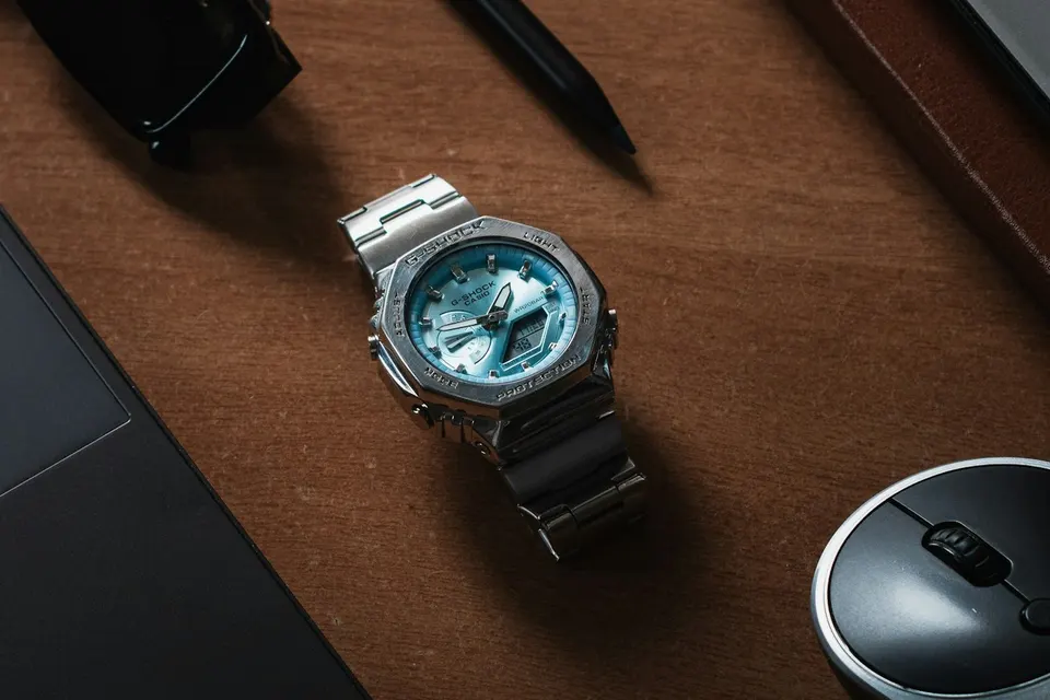 Vollmetall-G-Shock aus Edelstahl mit blauem Zifferblatt auf einem Schreibtisch zwischen Laptop, Stift und Sonnenbrille, gebürstetes Band und polierte Facetten sichtbar