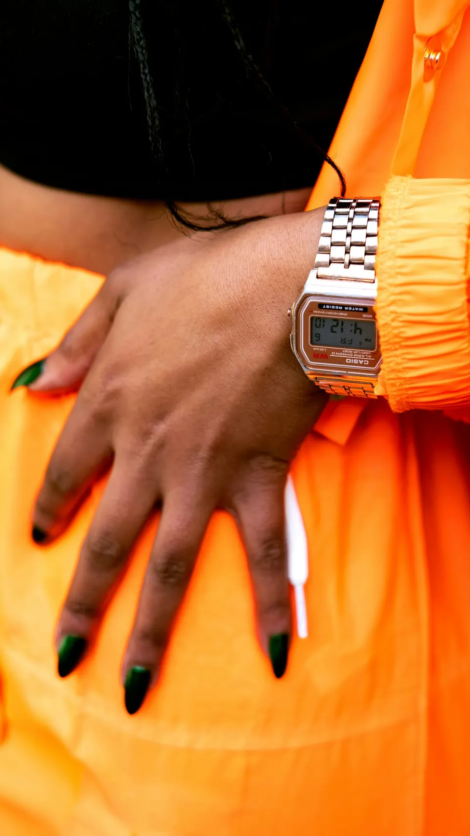 Frauenhand mit silberner Casio-Digitaluhr in Metall-Optik vor leuchtend orangefarbener Jacke und schwarzem Top