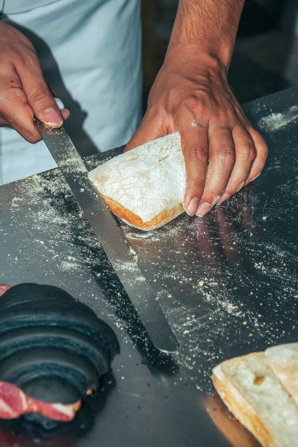 Ein Bäcker schneidet ein bemehltes Ciabatta mit einem langen Wellenschliffmesser auf einem Edelstahltisch