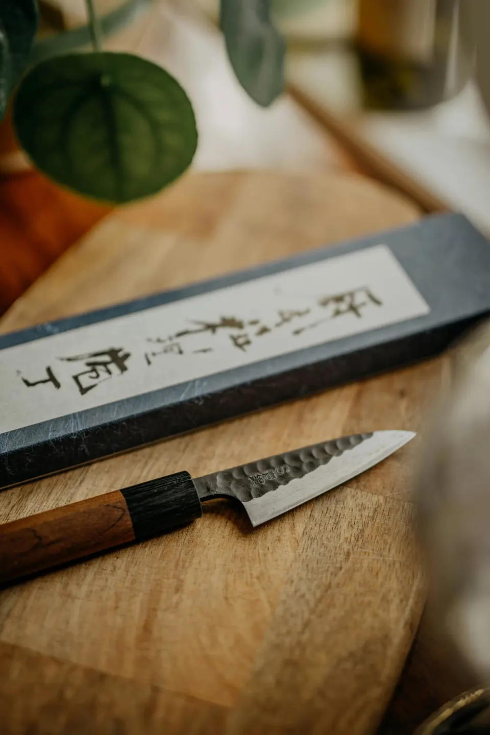 Ein kleines japanisches Messer mit gehämmerter Klingenflanke und Holzgriff liegt auf einem Holzbrett vor einer beschrifteten Papierbox