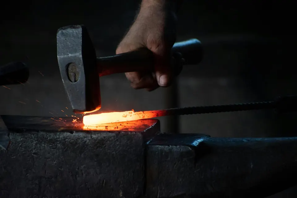 Eine Hand schlägt mit dem Hammer auf ein gelb glühendes Stück Stahl auf dem Amboss, Funken sprühen in einer dunklen Schmiede