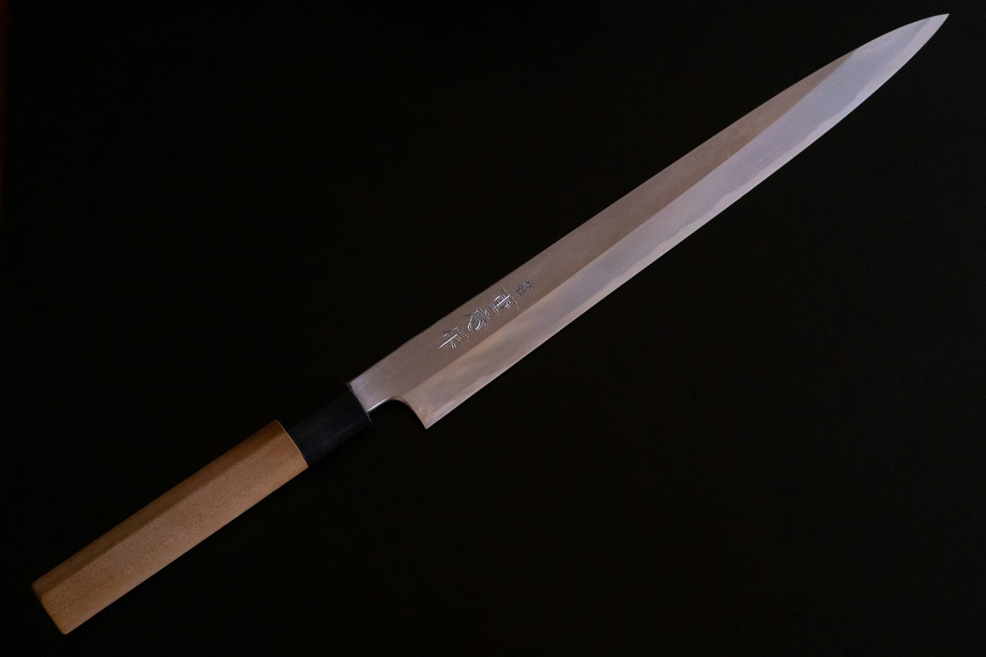 Ein langes japanisches Yanagiba mit hellem Holzgriff auf schwarzem Grund, die Grenze zwischen weichem Mantel und hartem Kern zieht als weiche Linie über die Klinge