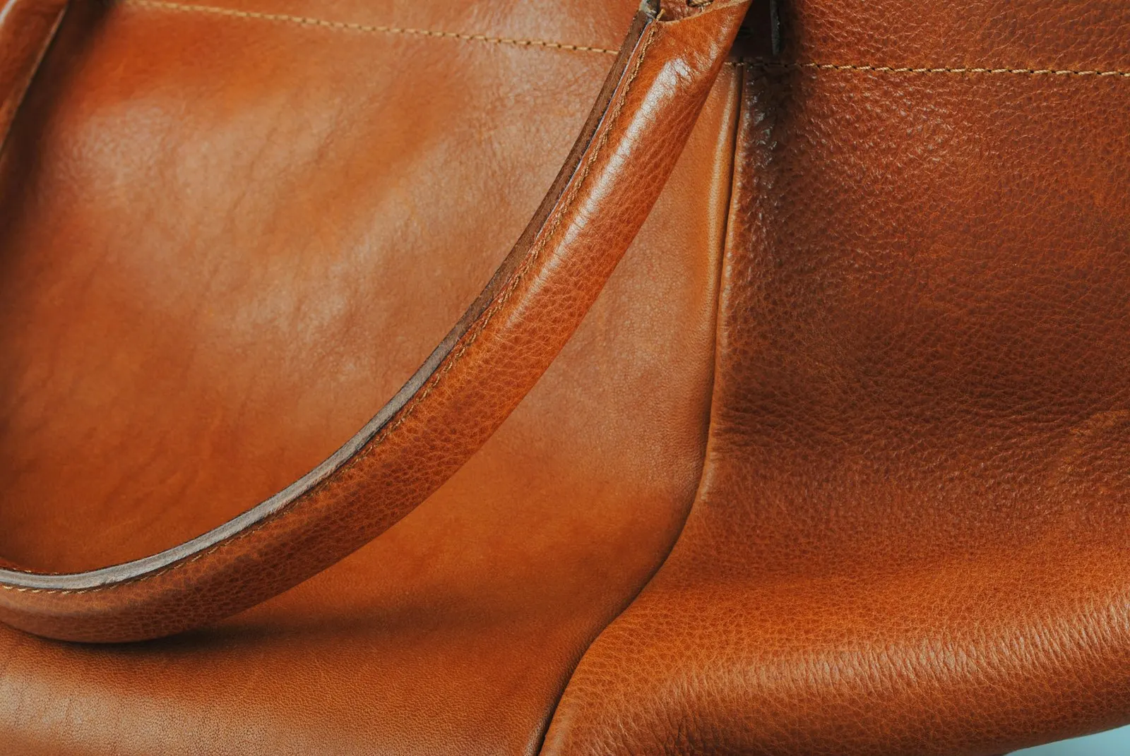 Nahaufnahme einer cognacfarbenen Ledertasche mit glatter, leicht glänzender Oberfläche, einem gerundeten Henkel und einer gleichmäßigen Steppnaht entlang der Kante