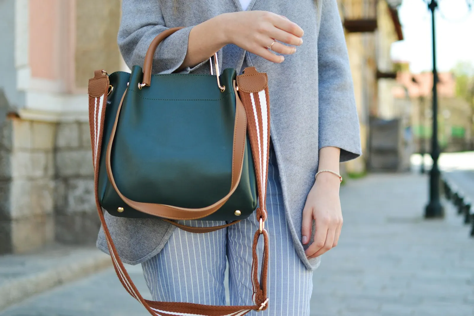 Dunkelgrüne Henkeltasche mit cognacfarbenen Henkeln, Metallfüßen am Boden und einem breiten, rot-weiß gestreiften Wechselriemen, getragen zu grauer Strickjacke und gestreifter Hose