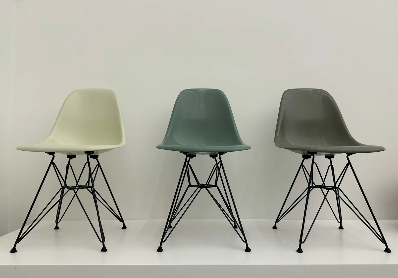 Drei Eames Plastic Side Chairs mit Drahtgestell in Creme, Graugrün und Grau vor weißer Wand im Vitra Design Museum