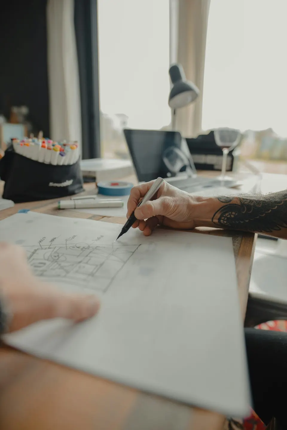 Hand zeichnet einen Grundriss mit Fineliner, im Hintergrund Marker und Schreibtischleuchte