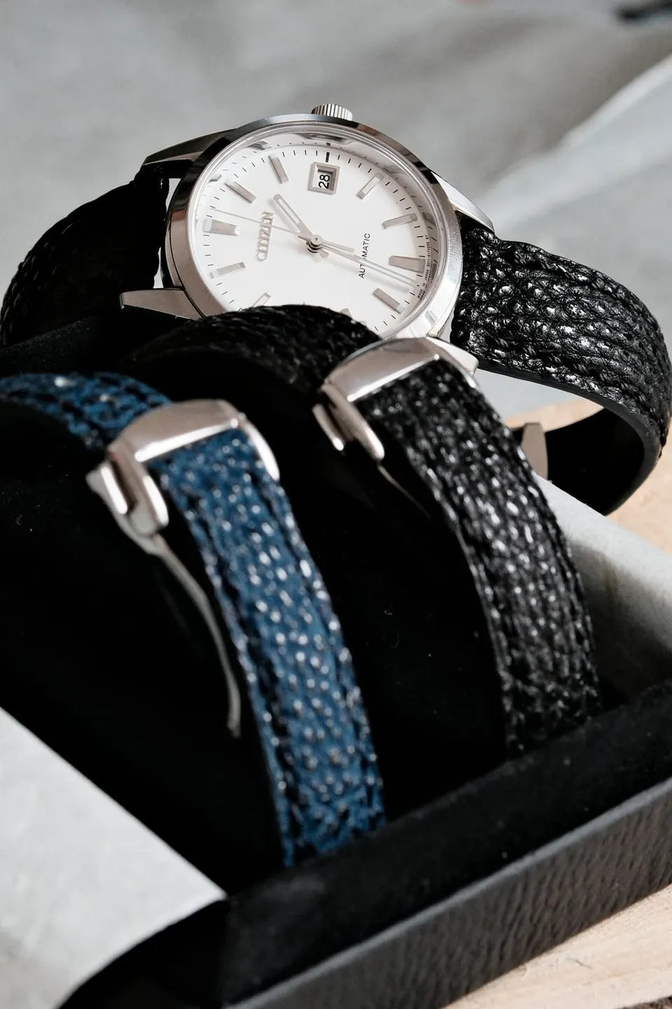 Automatikuhr in einem geöffneten Etui, daneben zwei Wechselbänder aus blauem Tweed und schwarzem genarbtem Material