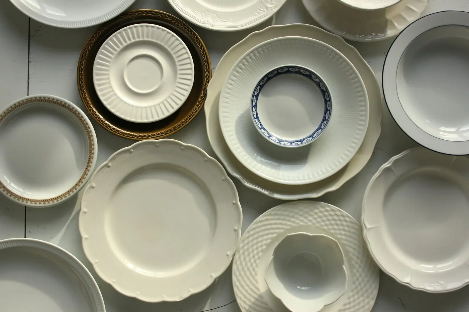 Viele weiße Teller und Schalen mit Reliefrändern, einige mit goldenem Rand, dicht nebeneinander von oben fotografiert