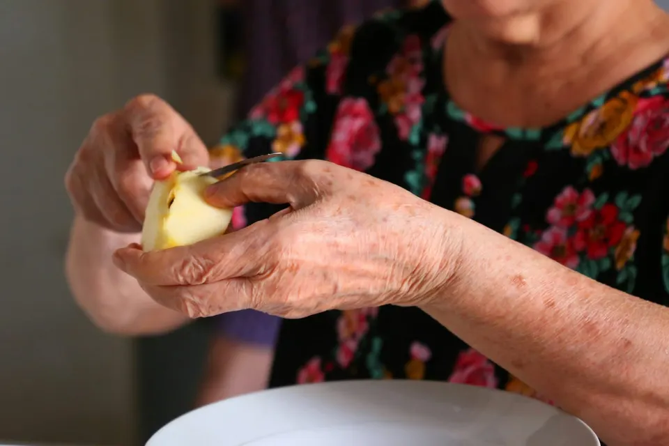 Eine ältere Frau schält einen Apfel mit einem kleinen Messer, die Hände nah an der Klinge
