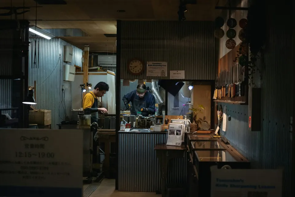 Blick in eine japanische Messerschleif-Werkstatt, zwei Handwerker an der Schleifstation, eine Wand mit Messern