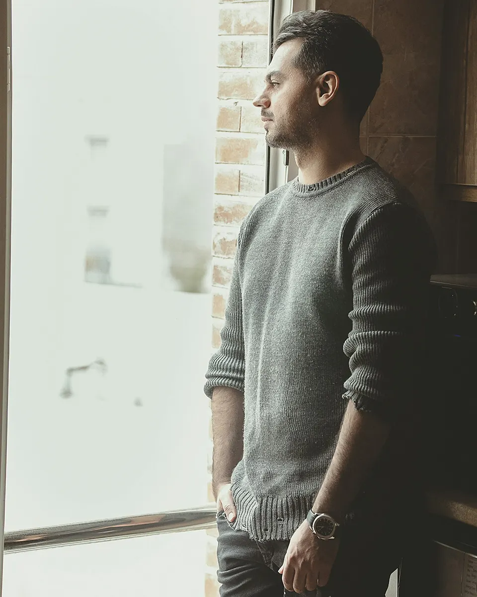 Mann in grauem Rundhals-Strickpullover mit Uhr lehnt am Fenster