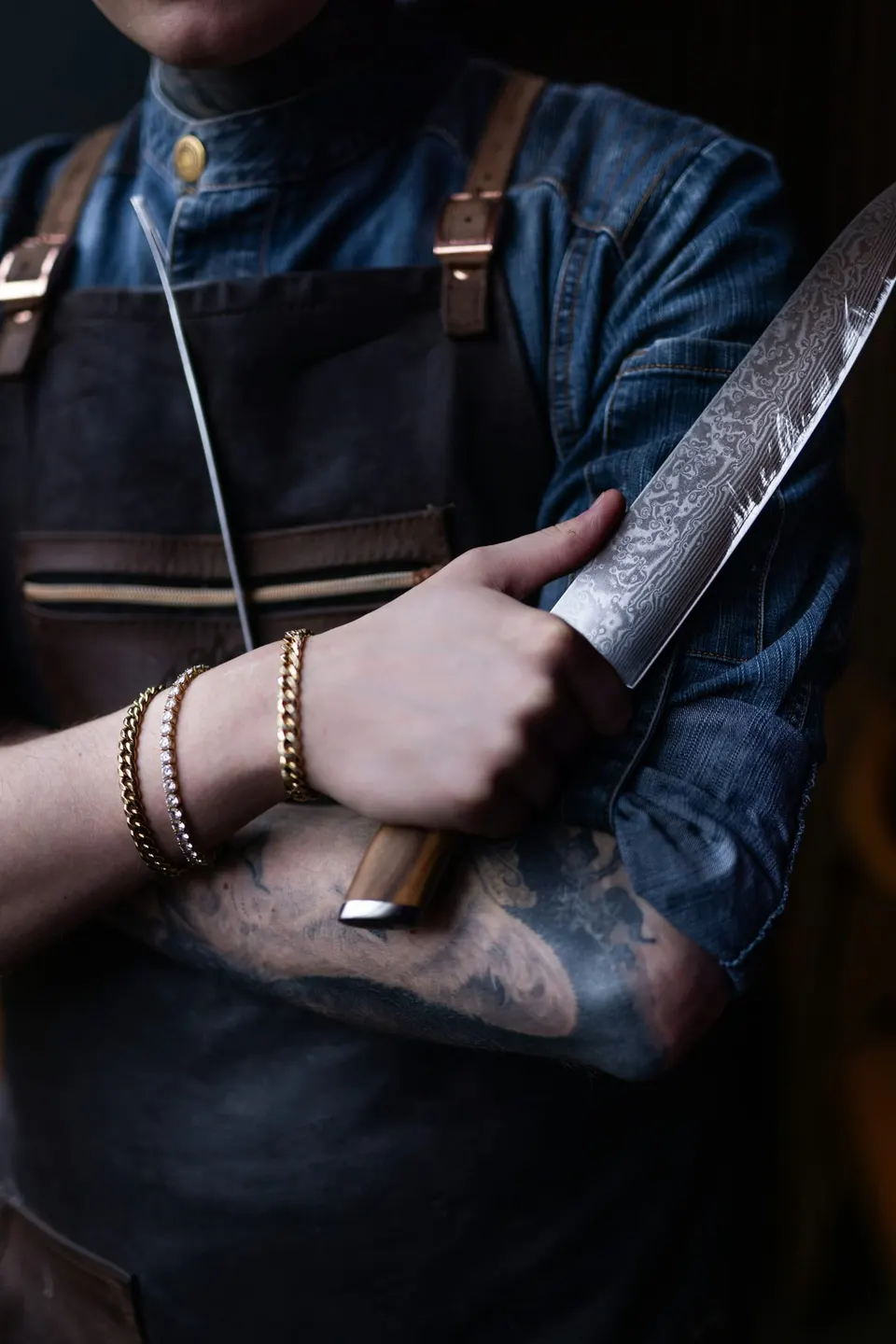 Ein Koch in Lederschürze hält ein Messer mit Holzgriff fest in der Faust, ein Wetzstahl klemmt unter dem Arm