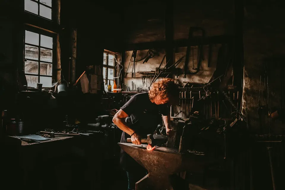 Ein Schmied schlägt in einer dunklen Werkstatt mit dem Hammer auf ein glühendes Werkstück auf dem Amboss