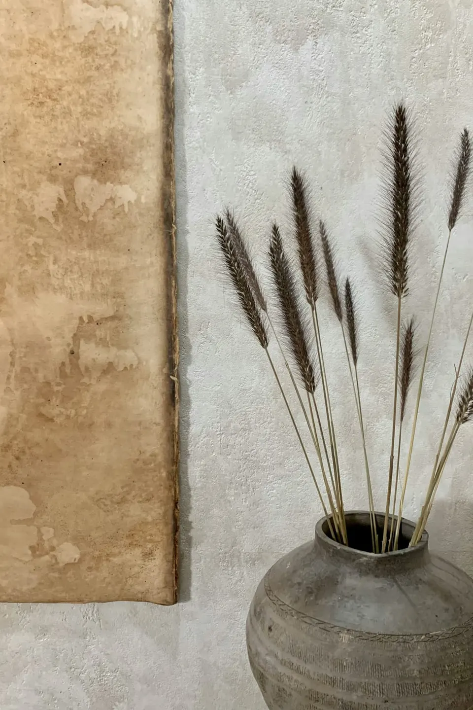 Graue Steingut-Krugvase mit Trockengräsern vor einer strukturierten Kalkputz-Wand in warmen Beigetönen