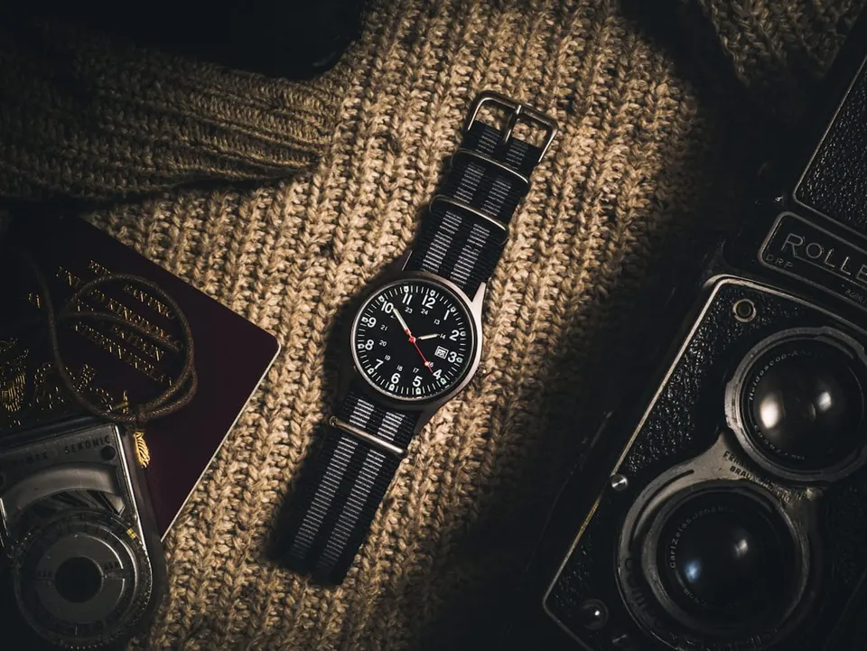 Feldfoermige Uhr mit Textilband liegt auf grobem Strick neben alter Rollfilmkamera und Reisepass